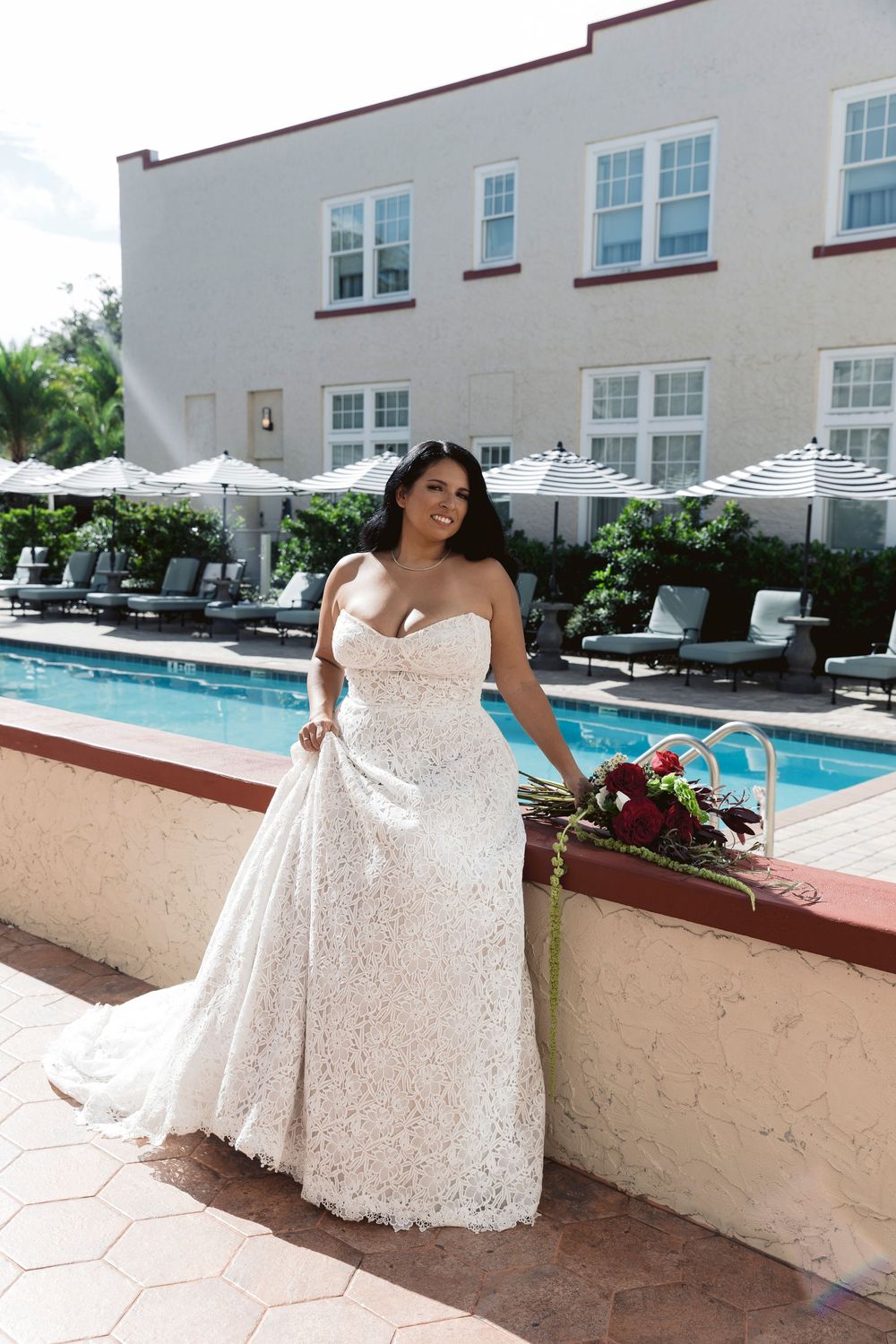 Radiant bride in strapless lace wedding gown poses poolside holding a deep red floral bouquet outdoors.