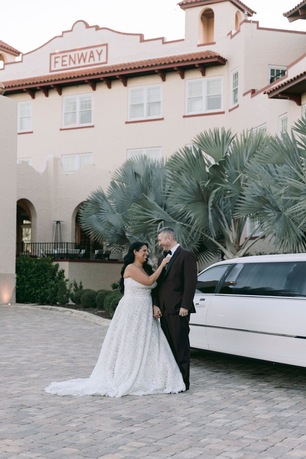 Bride and groom pose beside a white limousine outside the elegant Fenway building, surrounded by palm trees.