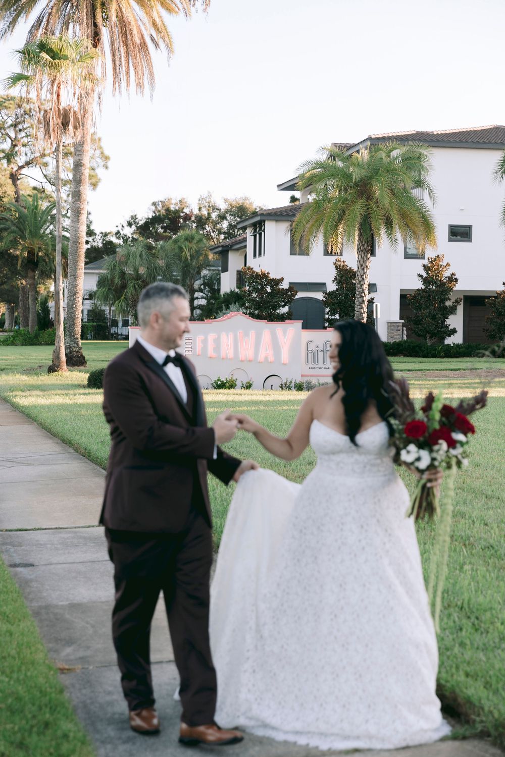 Bride and groom hold hands on a palm-lined sidewalk near a Fenway sign, bride holding red bouquet in white gown.
