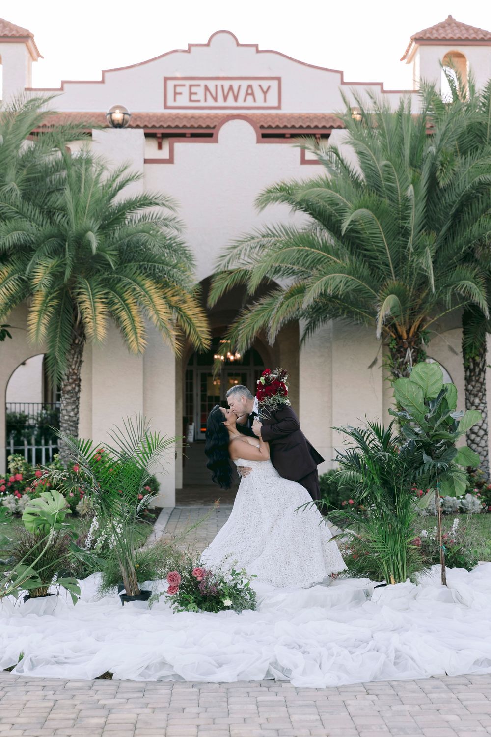 Bride and groom share a romantic dip kiss in front of the Fenway building surrounded by tropical palms.