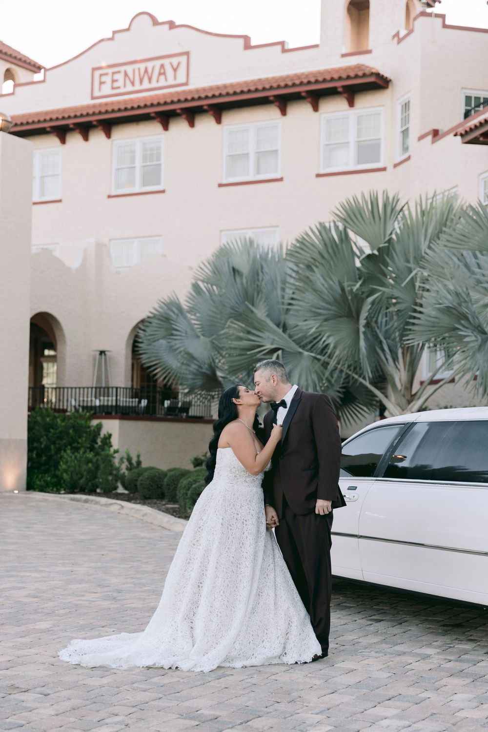 Bride and groom share a kiss outside the Fenway venue beside a white limo, surrounded by palm trees.