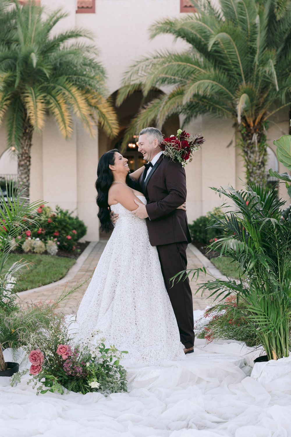 Bride and groom embrace outdoors surrounded by tropical palm trees and floral arrangements in a romantic wedding portrait.