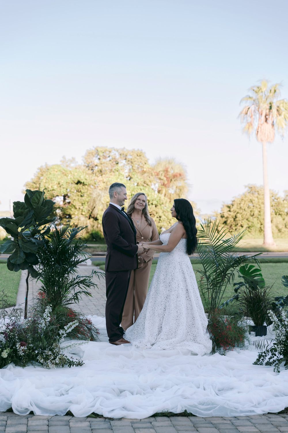 Bride and groom exchange vows with officiant at outdoor wedding ceremony surrounded by tropical greenery and palm trees.
