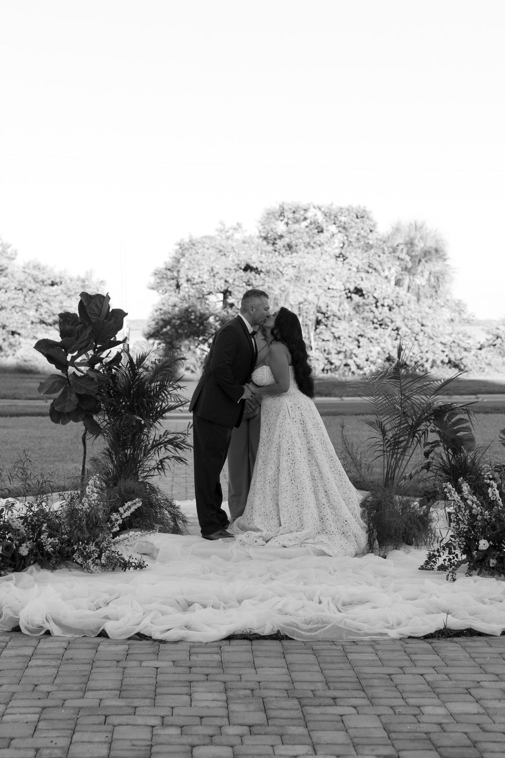 Black and white photo of a bride and groom sharing a kiss at an outdoor wedding ceremony with floral arrangements.