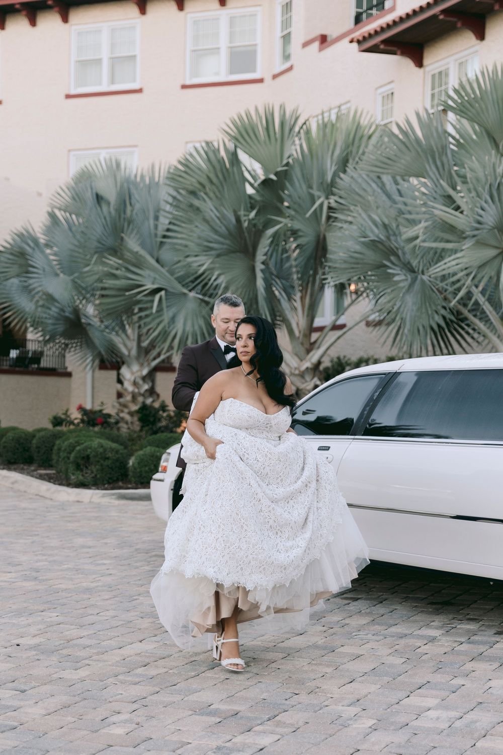 Bride and groom walking beside a white limousine with palm trees and a Mediterranean-style building in the background.