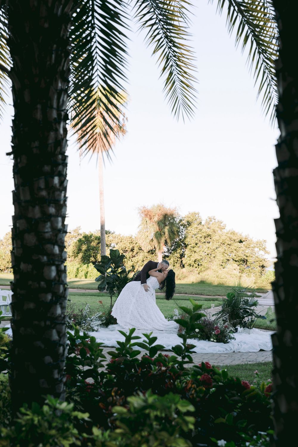 Bride and groom share a romantic dip kiss surrounded by lush palm trees and tropical gardens at sunset.