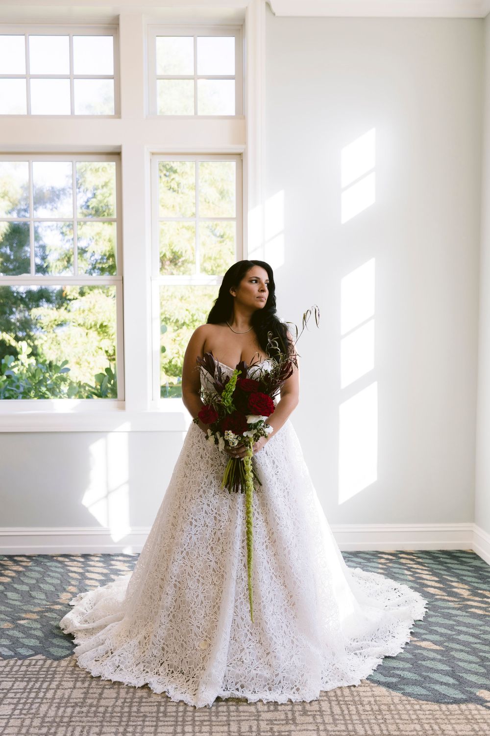Elegant bride in strapless lace ball gown holds red bouquet while posing in sunlit room with large windows.