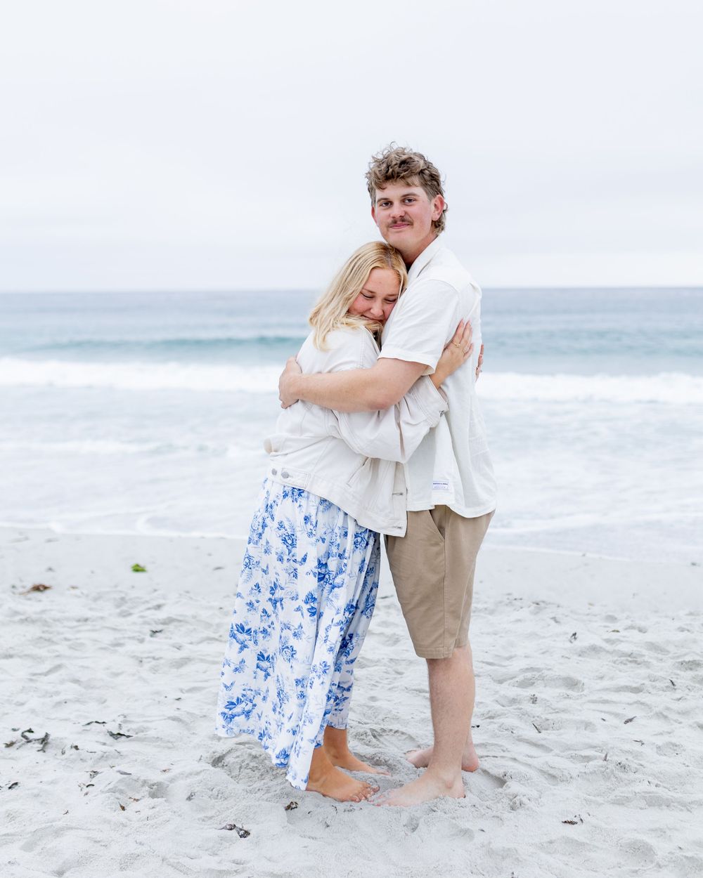 A couple embraces on a sandy beach, woman in floral skirt and man in white shirt, with ocean waves in the background.