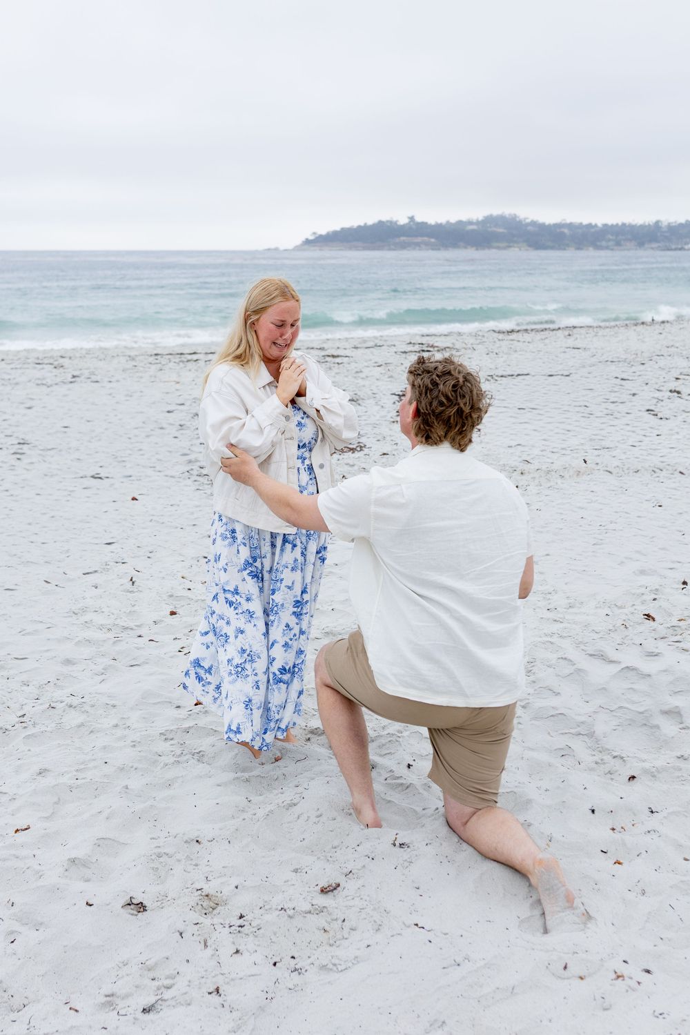 Man proposes on one knee to woman on sandy beach, ocean waves in background, overcast sky.