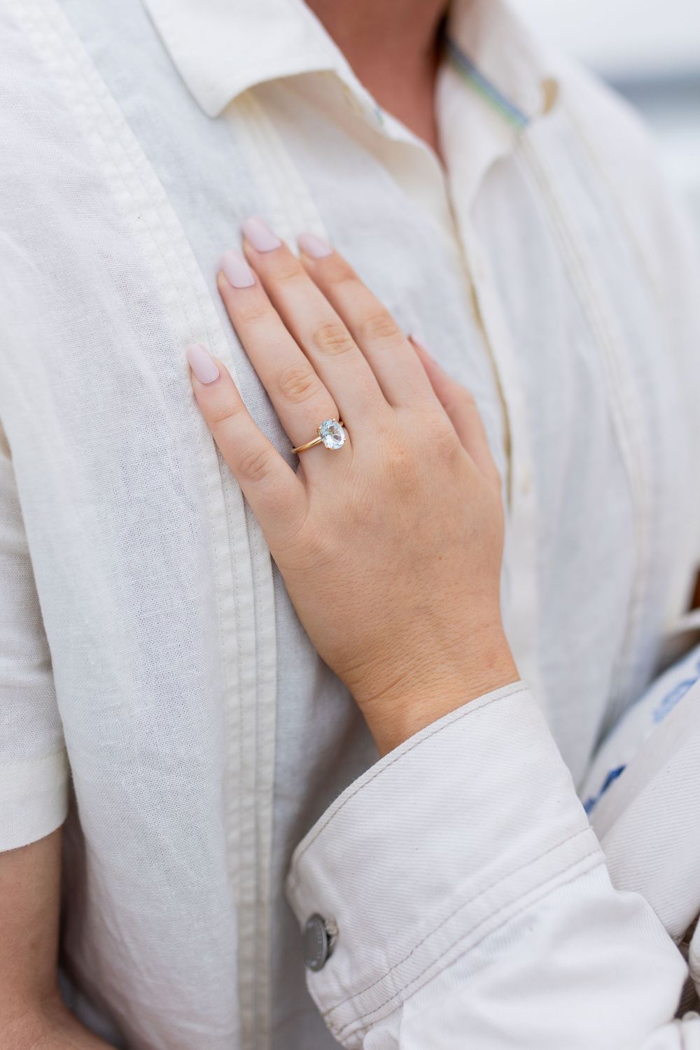 Close-up of a woman's hand wearing a delicate gold ring with a light blue gemstone, resting on a white linen shirt.