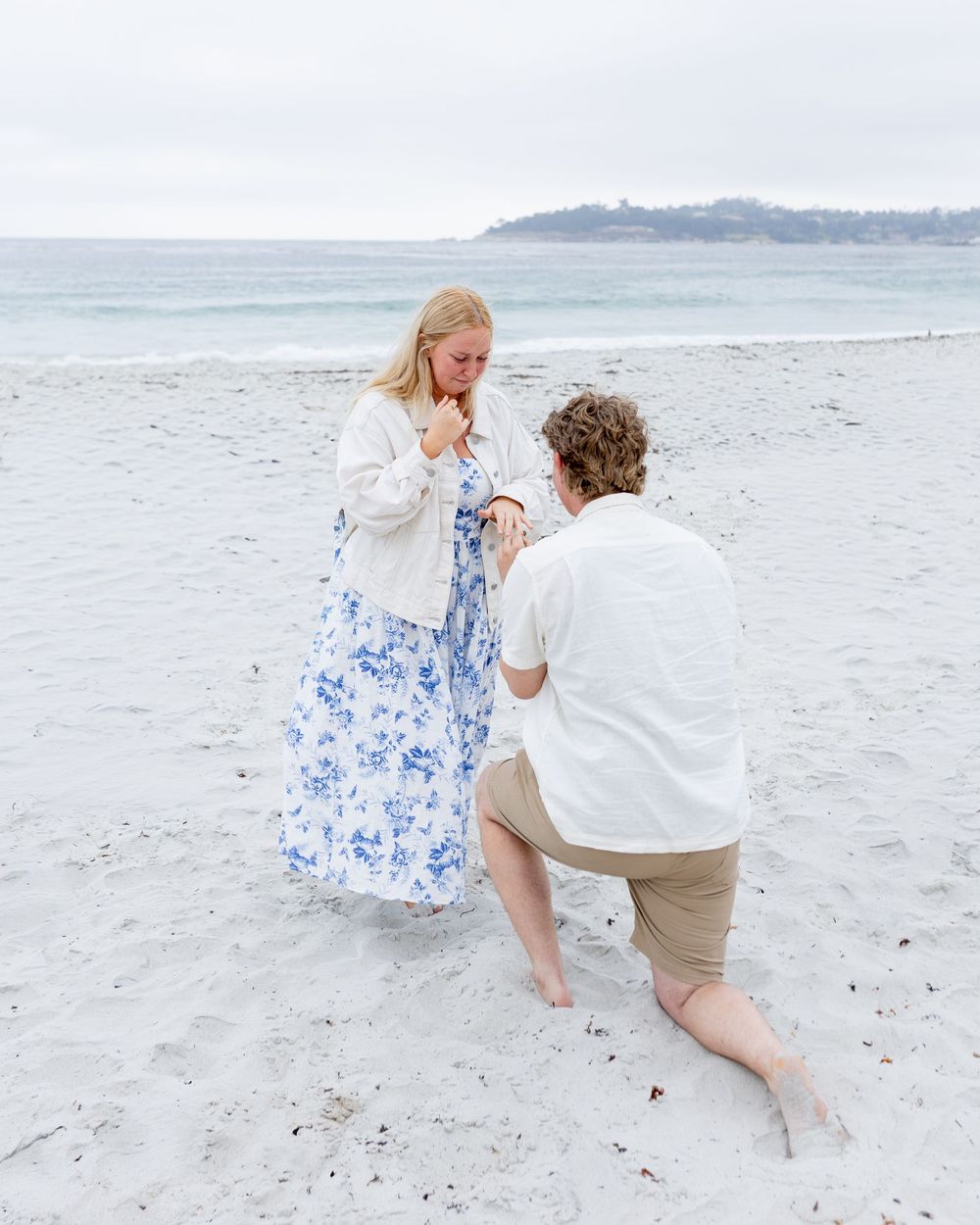 Man proposes on bended knee to woman in blue floral dress on a sandy beach by the ocean.