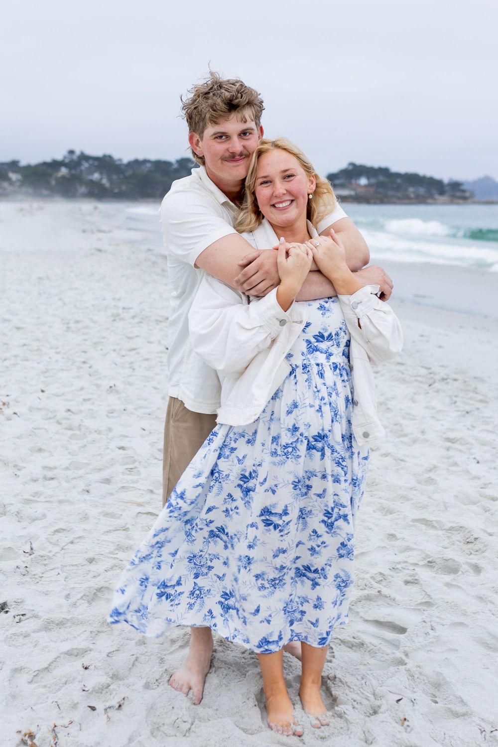 A couple embraces on a sandy beach, the woman wearing a blue floral dress, both smiling joyfully with ocean waves behind them.