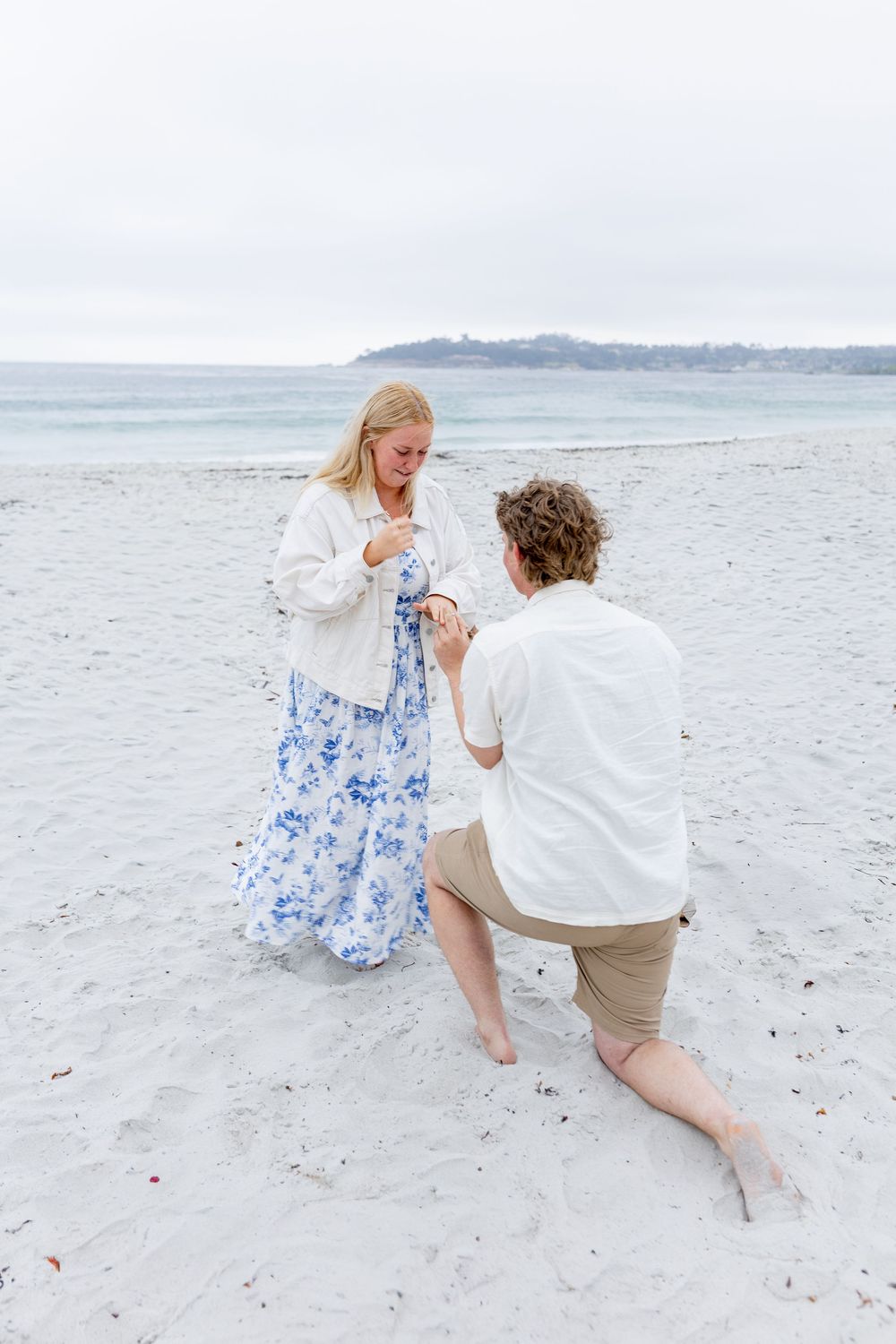 Man proposing on one knee to woman in floral dress on a sandy beach with ocean backdrop.