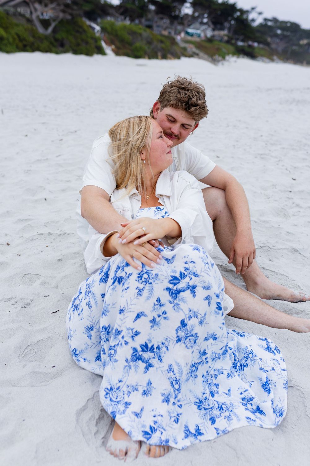 Couple sitting on white sand beach, woman in blue floral dress leaning against man in white shirt, smiling together.