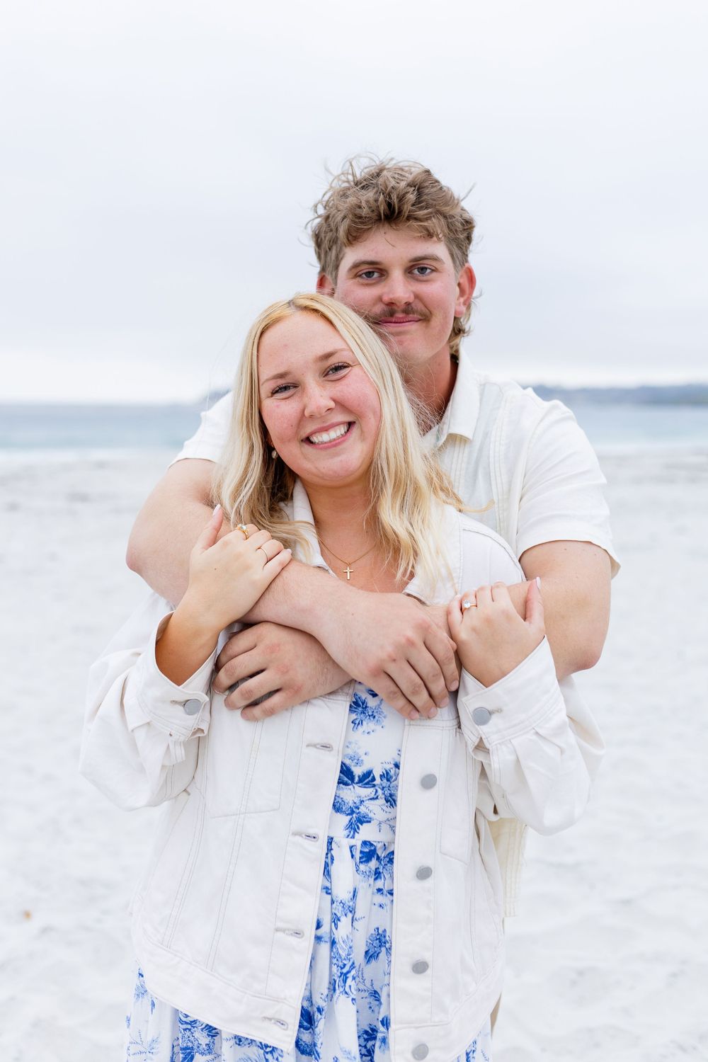 A smiling couple embraces on a sandy beach, woman in white denim jacket and blue floral dress, man in white shirt behind her.