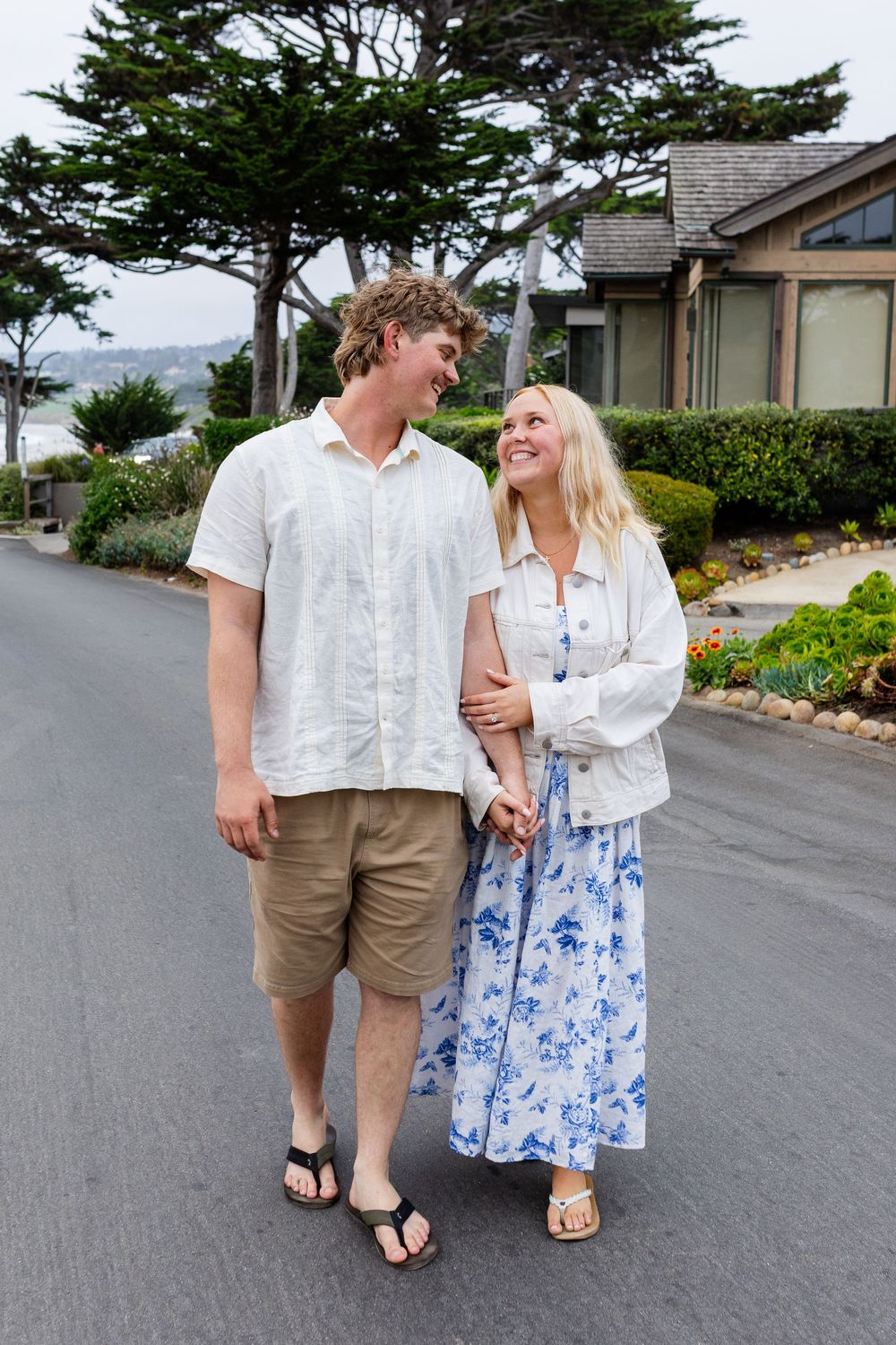 A young couple walks hand-in-hand down a quiet residential street, smiling at each other on a cloudy day.