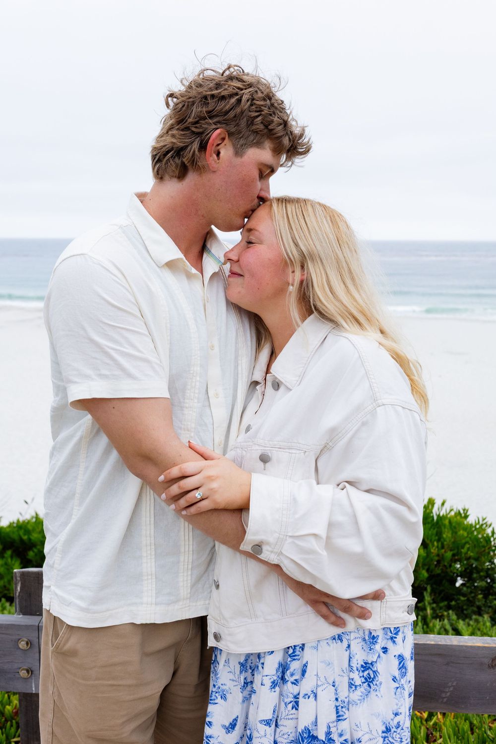 Couple sharing a tender forehead kiss while embracing near the ocean, dressed in white and floral.