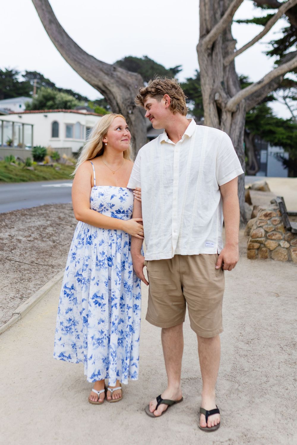 A young couple in summer attire walks together outdoors near a large tree, smiling at each other.