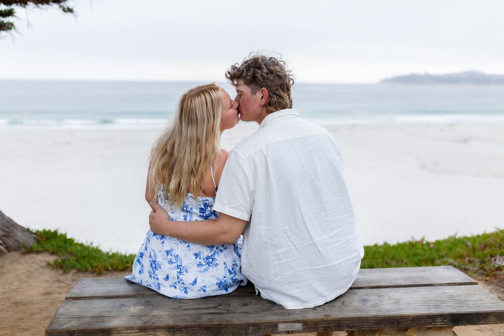 Couple sharing a kiss on a bench overlooking a serene beach with ocean views and cloudy skies.