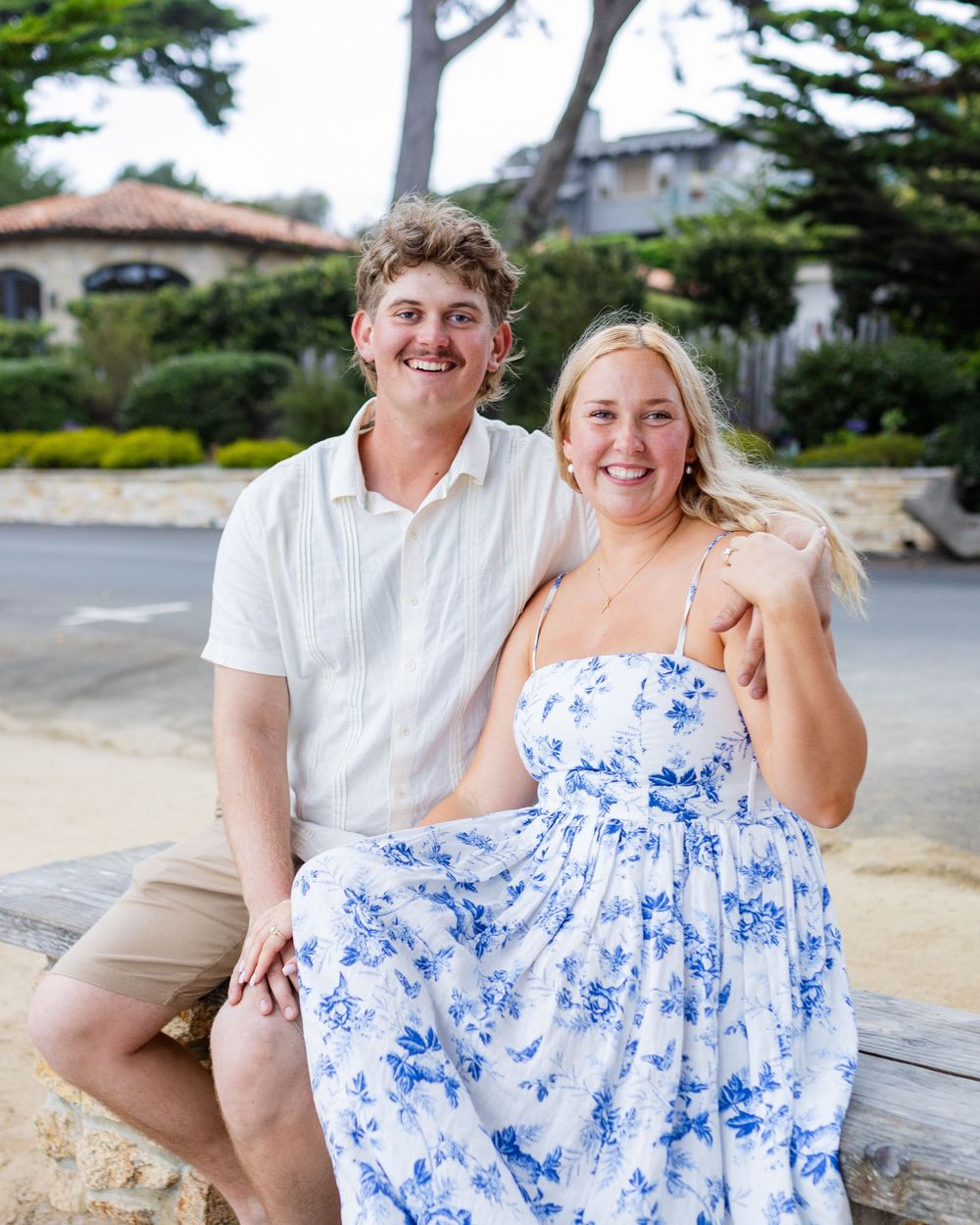 A smiling couple poses outdoors on a bench, man in white shirt and woman in blue floral sundress, with greenery behind them.