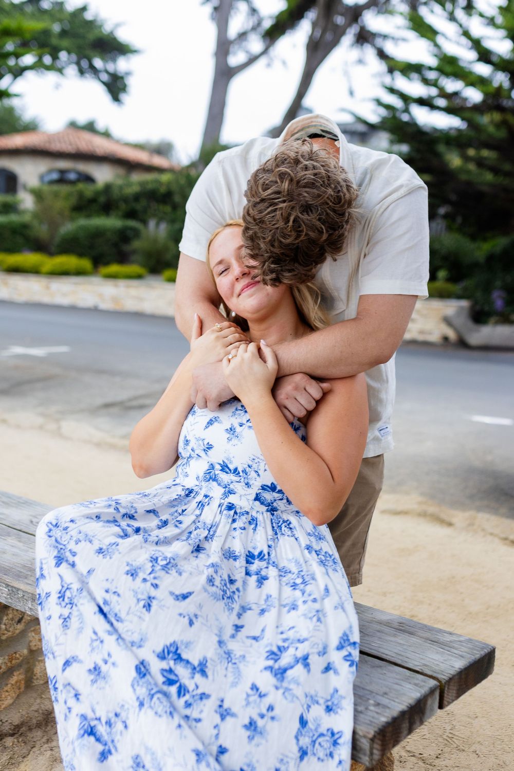 A couple sharing a tender embrace outdoors, woman in blue floral dress seated on bench while man hugs her from behind.