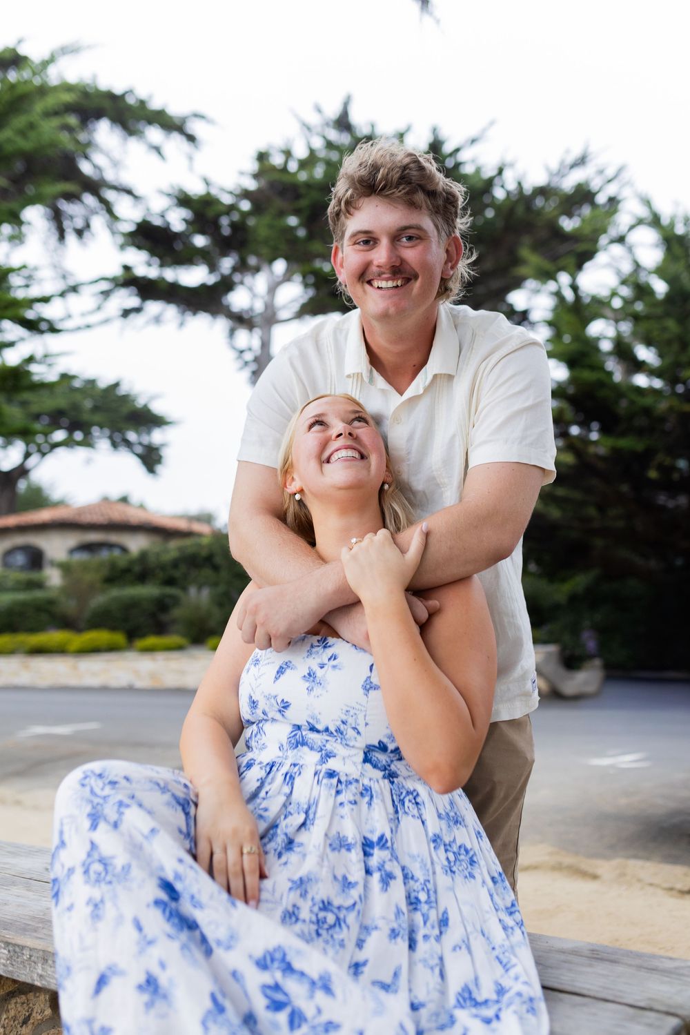 A smiling couple poses outdoors, man in white polo embracing woman in blue floral dress, with green trees behind them.