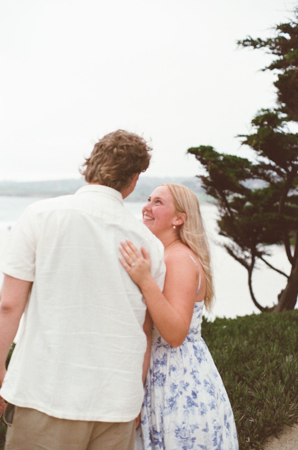 Couple sharing a romantic moment outdoors near the ocean, woman in floral dress smiling up at man in white linen shirt.