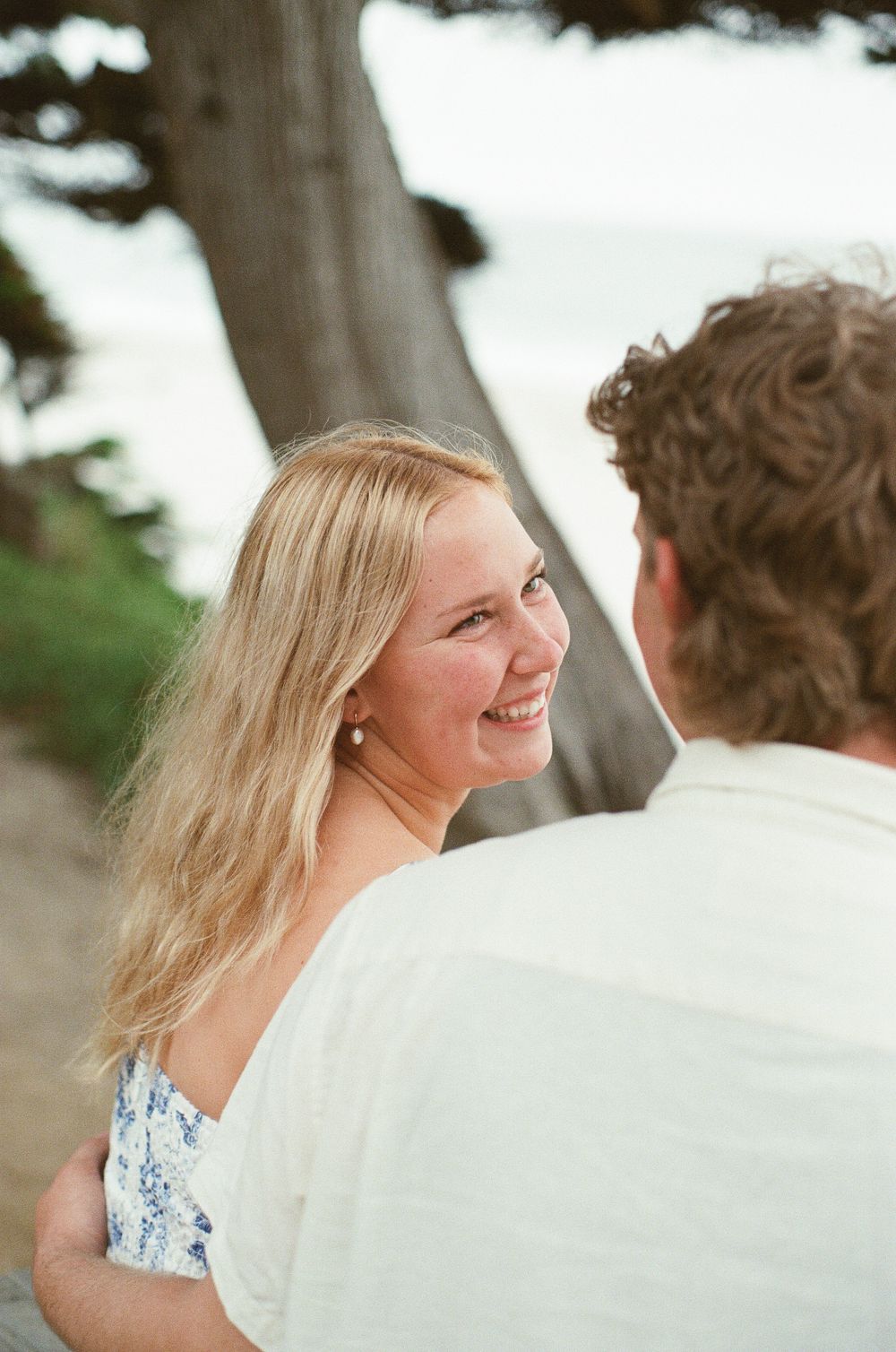 A smiling blonde woman in a floral dress looks up at a man near a beach tree.