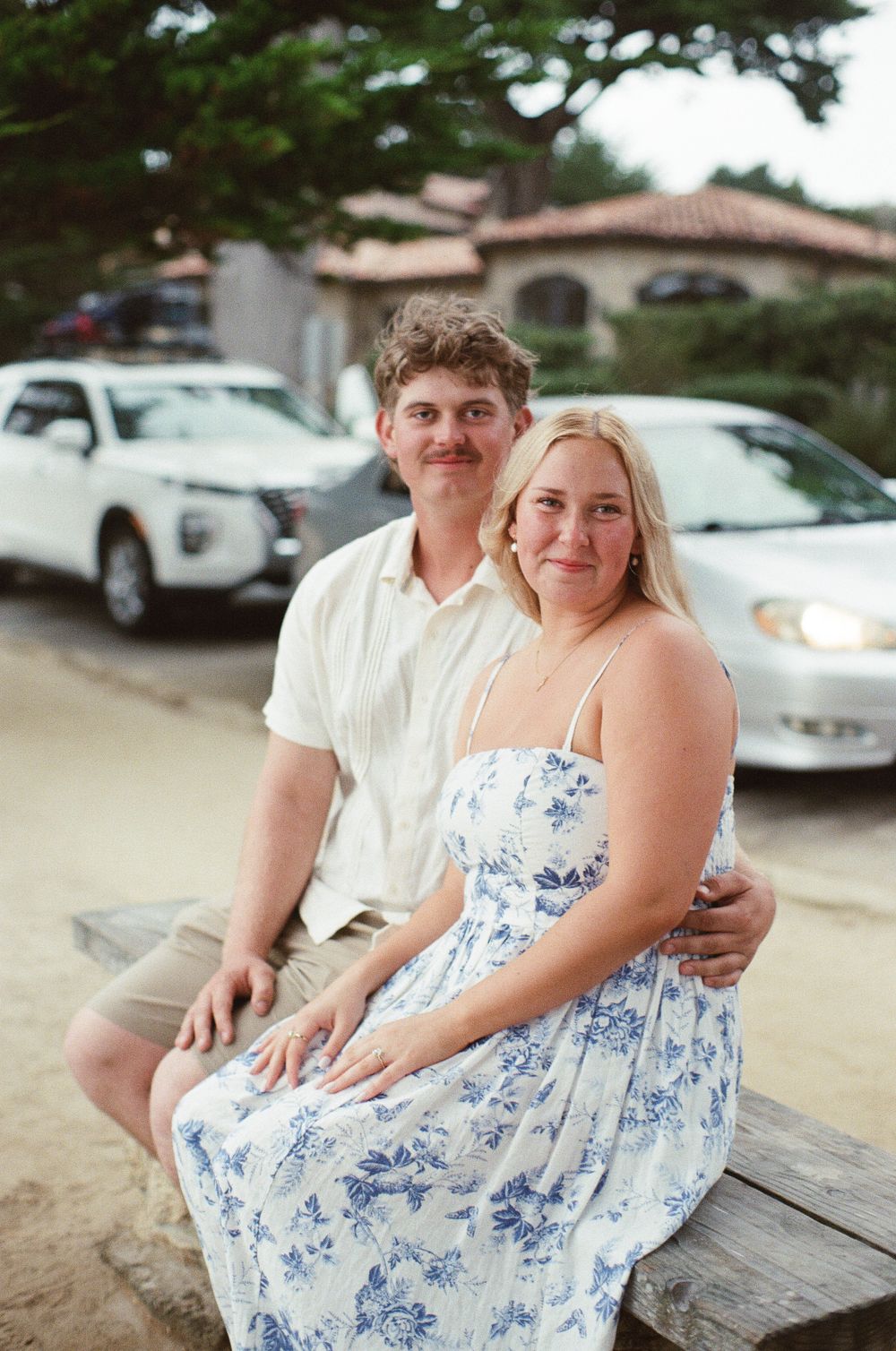 A smiling couple sits together outdoors near parked cars, woman in floral dress, man in white shirt.
