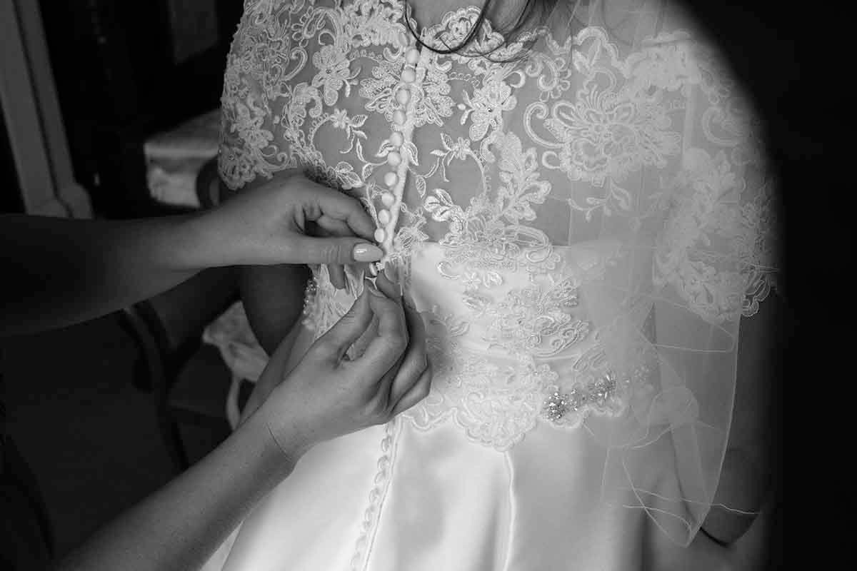 Black and white photo of a bride getting her lace wedding dress buttoned up by a bridesmaid at Blaithwaite House