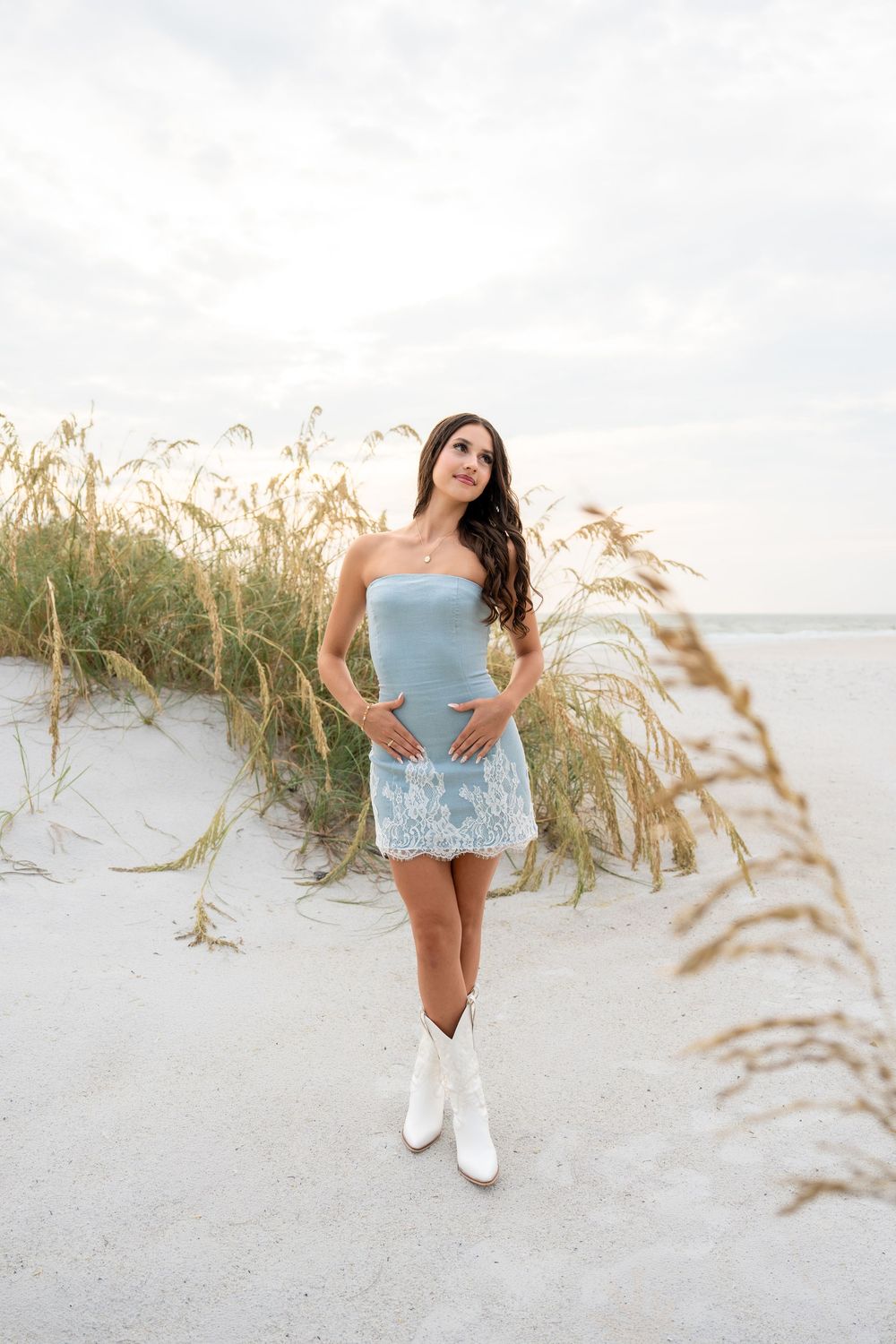Young brunette woman in blue strapless mini dress and white cowboy boots poses among sea oats on sandy beach.