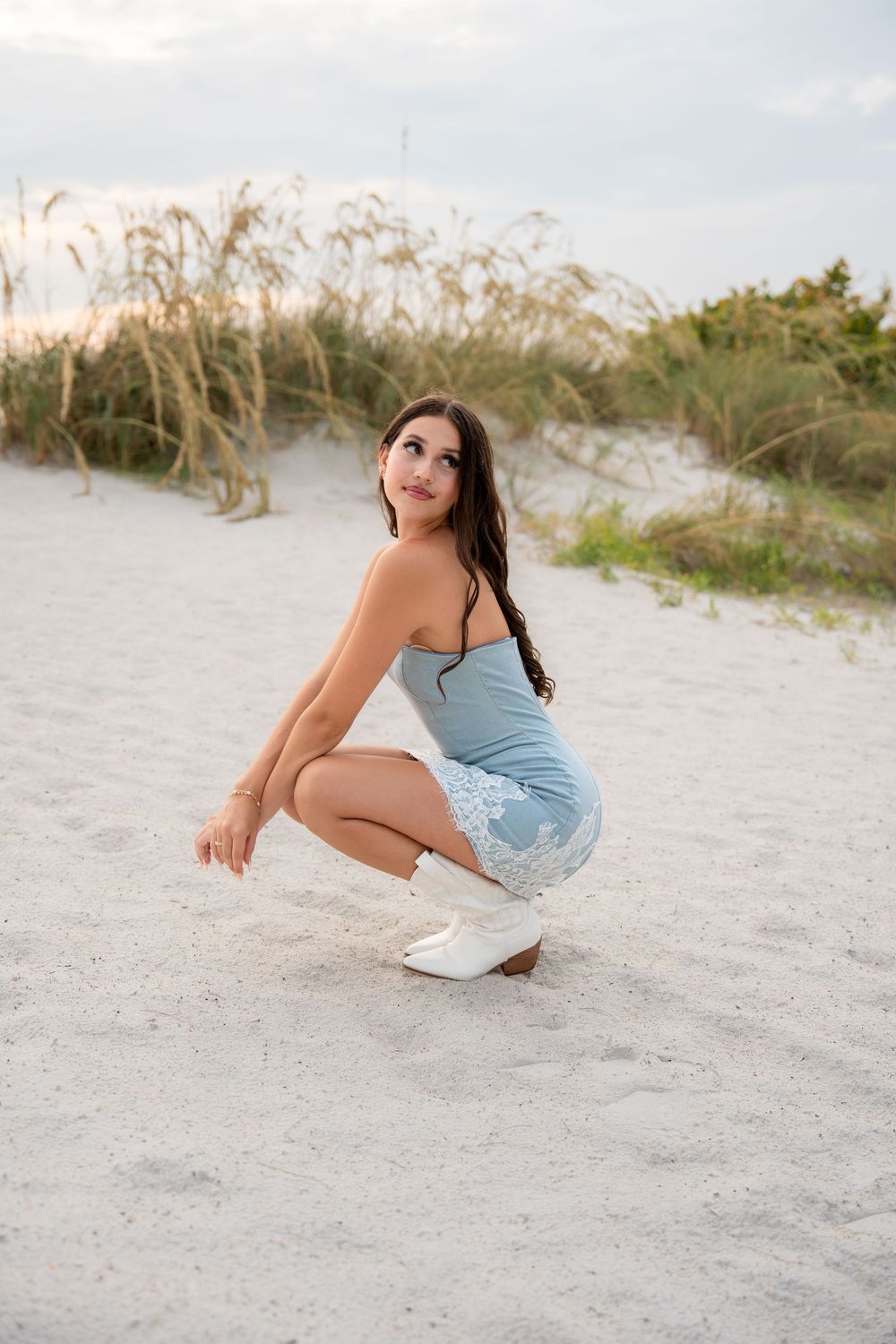 Young brunette woman in blue lace strapless mini dress crouches on white sand beach dunes at golden hour.