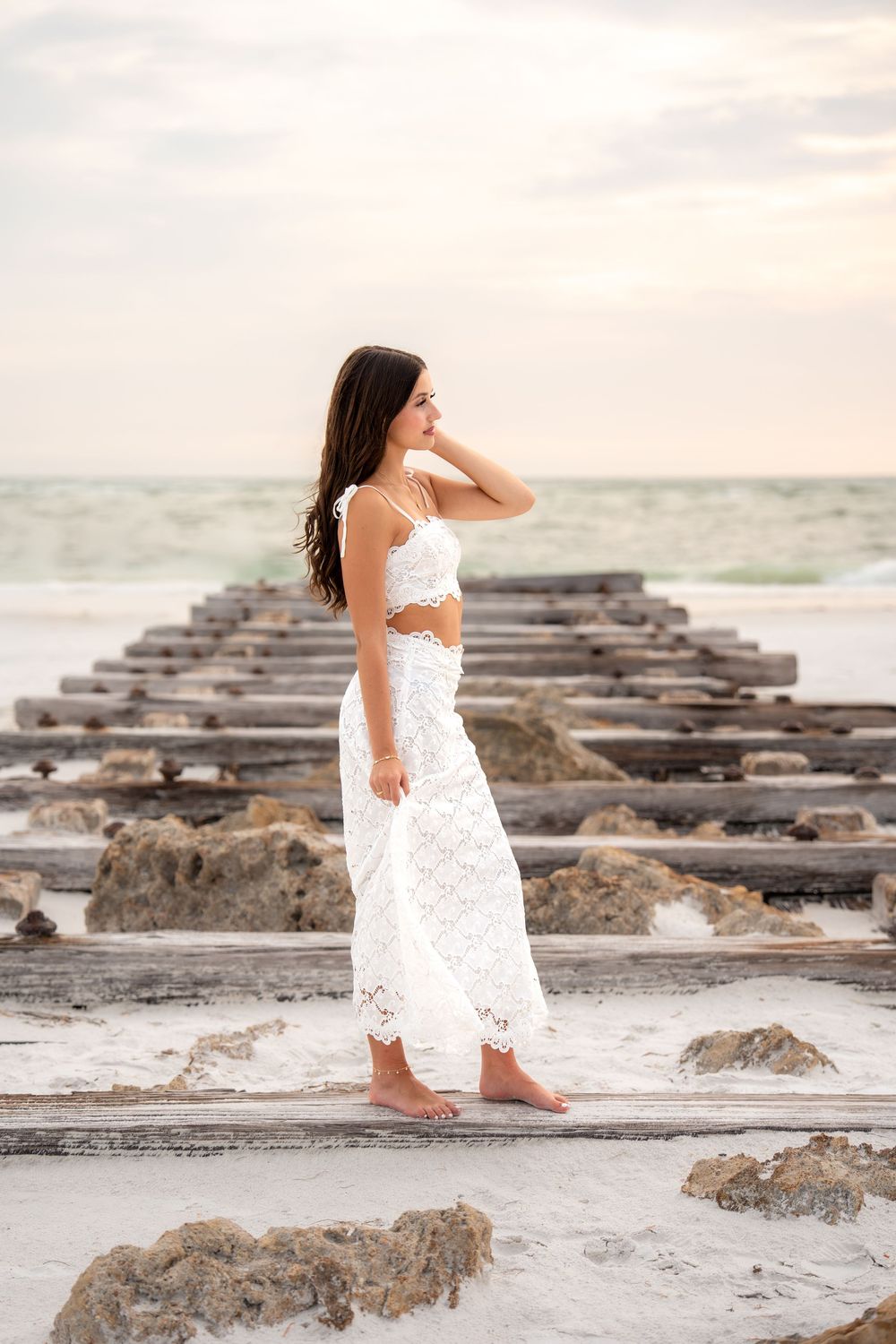 Young brunette woman in white lace two-piece outfit stands on rocky beach ruins with ocean waves at sunset.