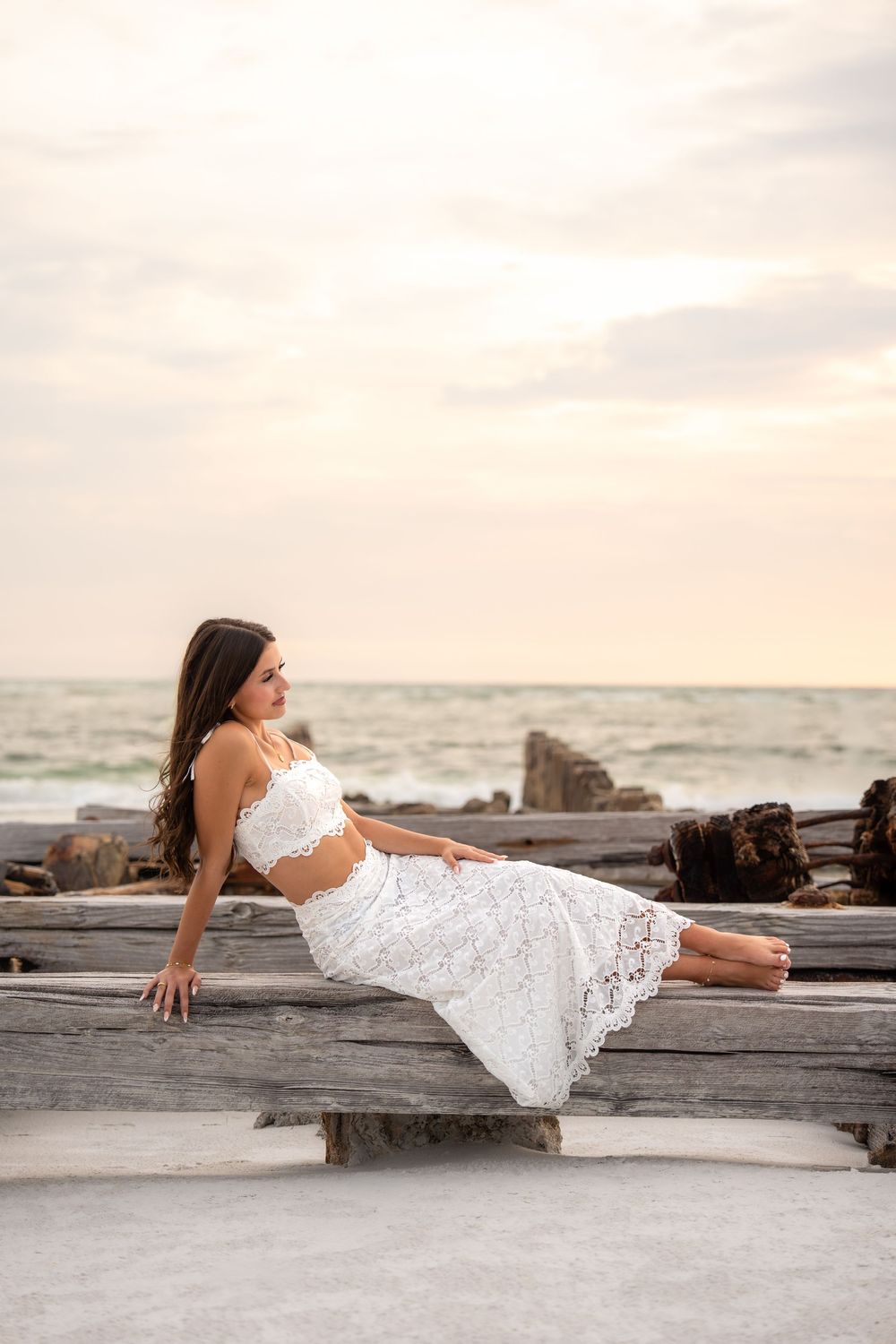 Woman in white lace crop top and skirt poses on weathered driftwood with ocean backdrop at dusk.