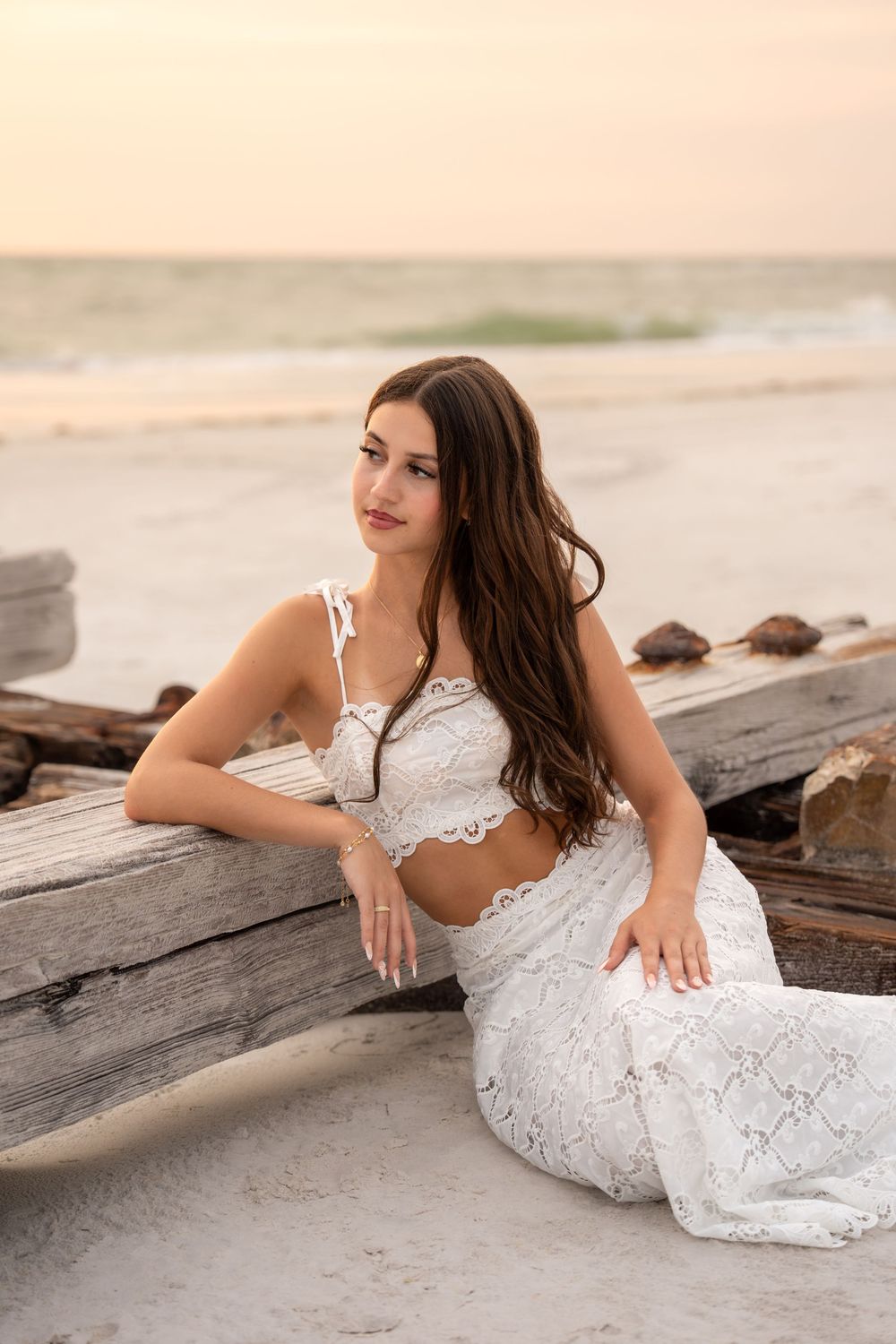 Elegant brunette in white lace two-piece sits against driftwood on a peaceful beach at sunset.