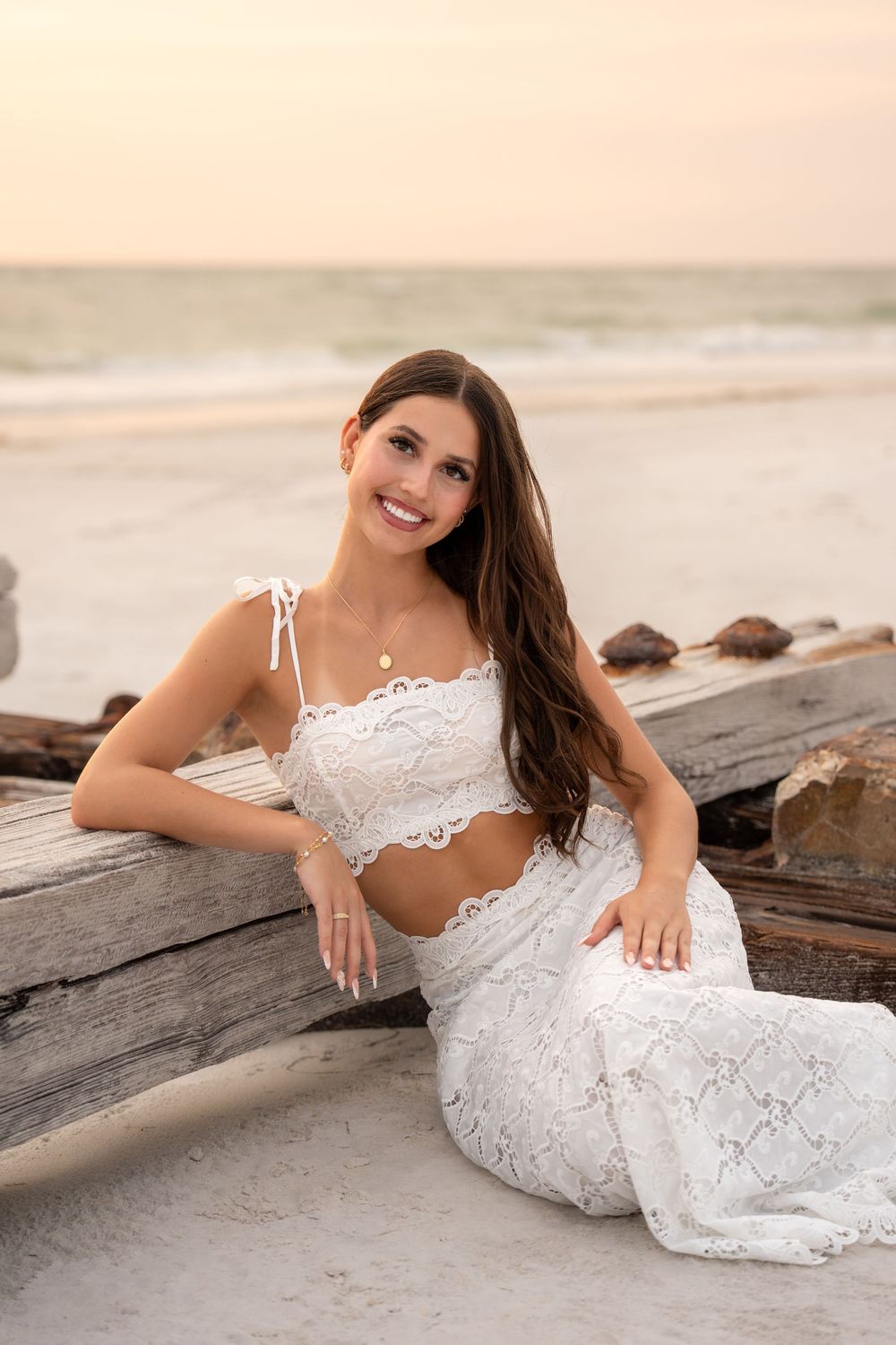 Smiling woman in white lace outfit leans against driftwood on a sandy beach during golden sunset.