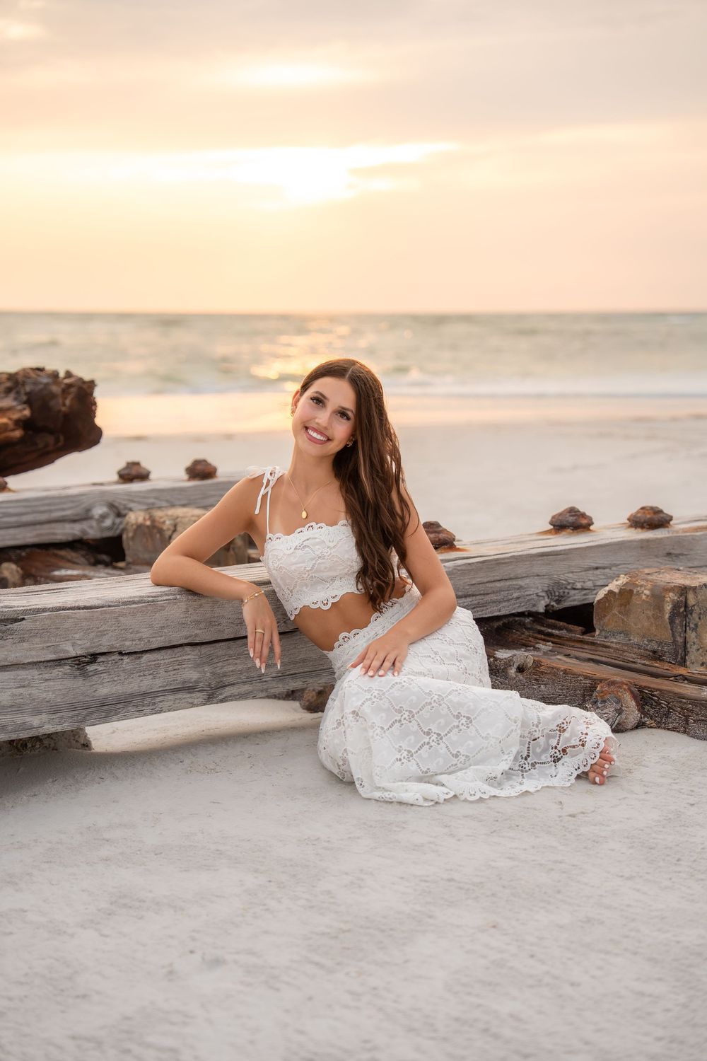 Happy brunette woman in white lace set sits on sand near driftwood during a pastel beach sunset.