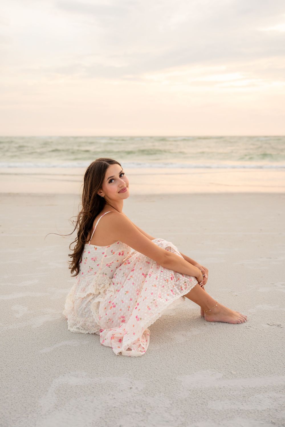 Brunette woman in floral sundress sits barefoot on white sand beach during a pastel sunset.