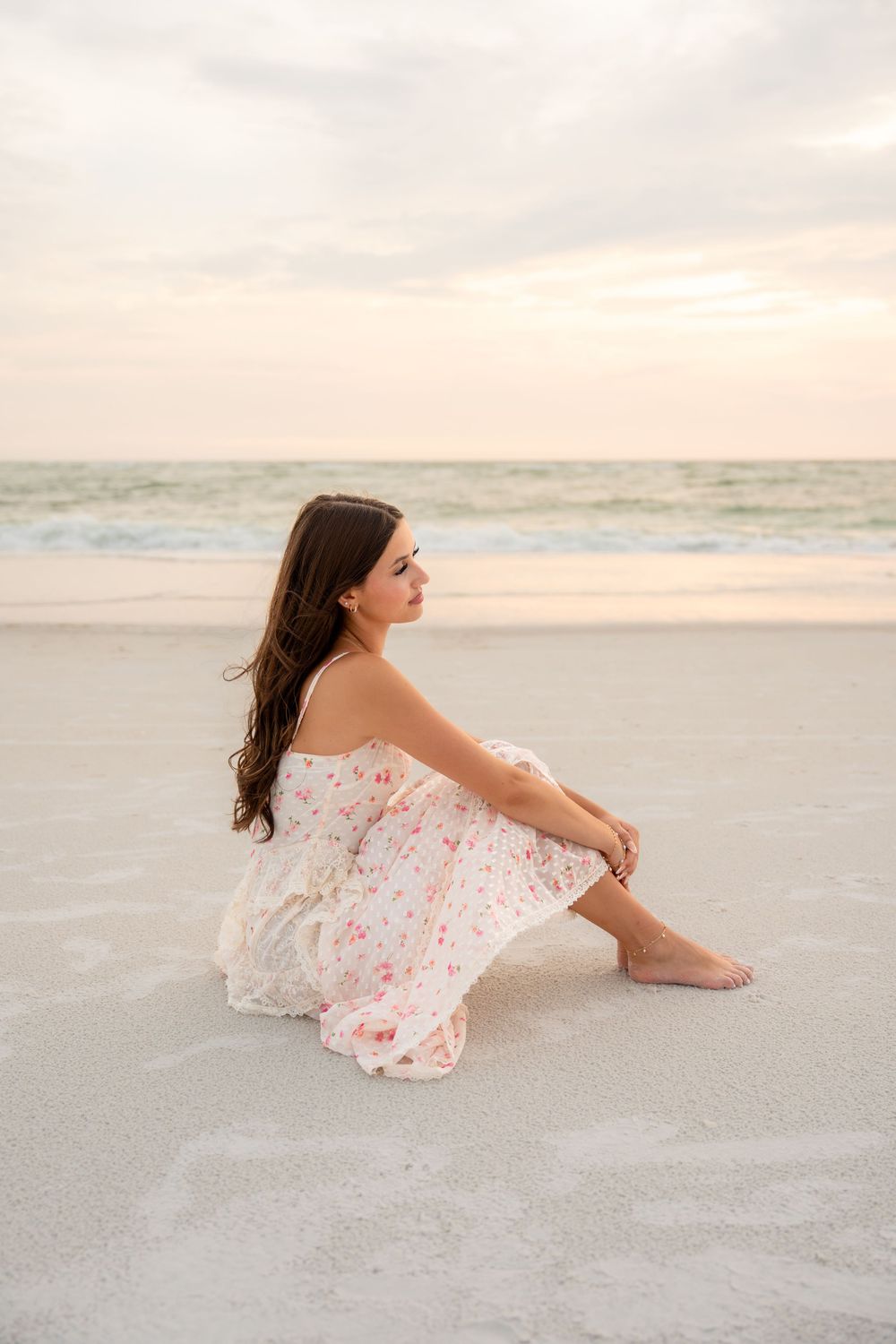 Young woman in floral dress sits gazing at the ocean on a serene white sand beach at sunset.