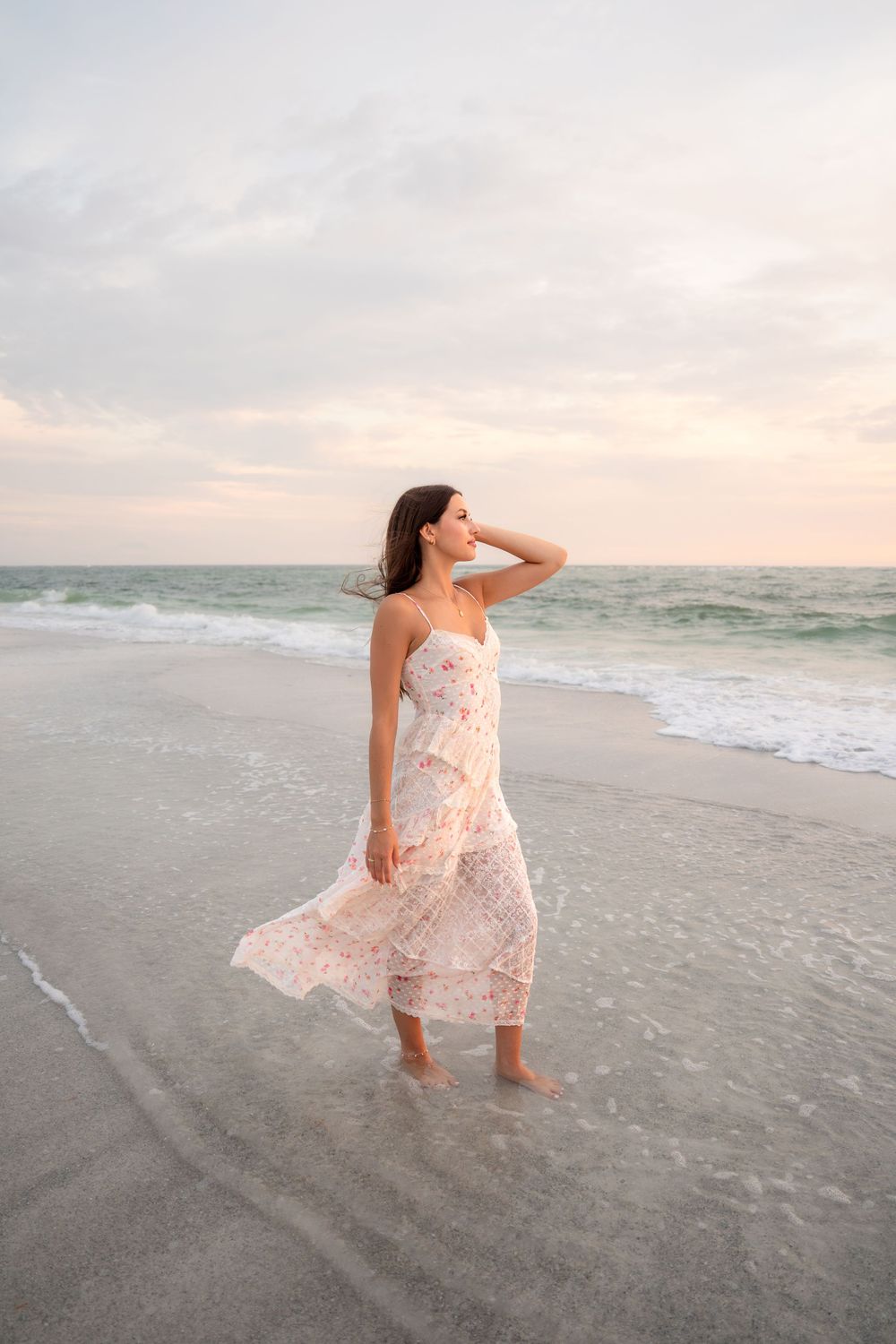 Barefoot woman in white floral dress standing in ocean waves at beach sunset.