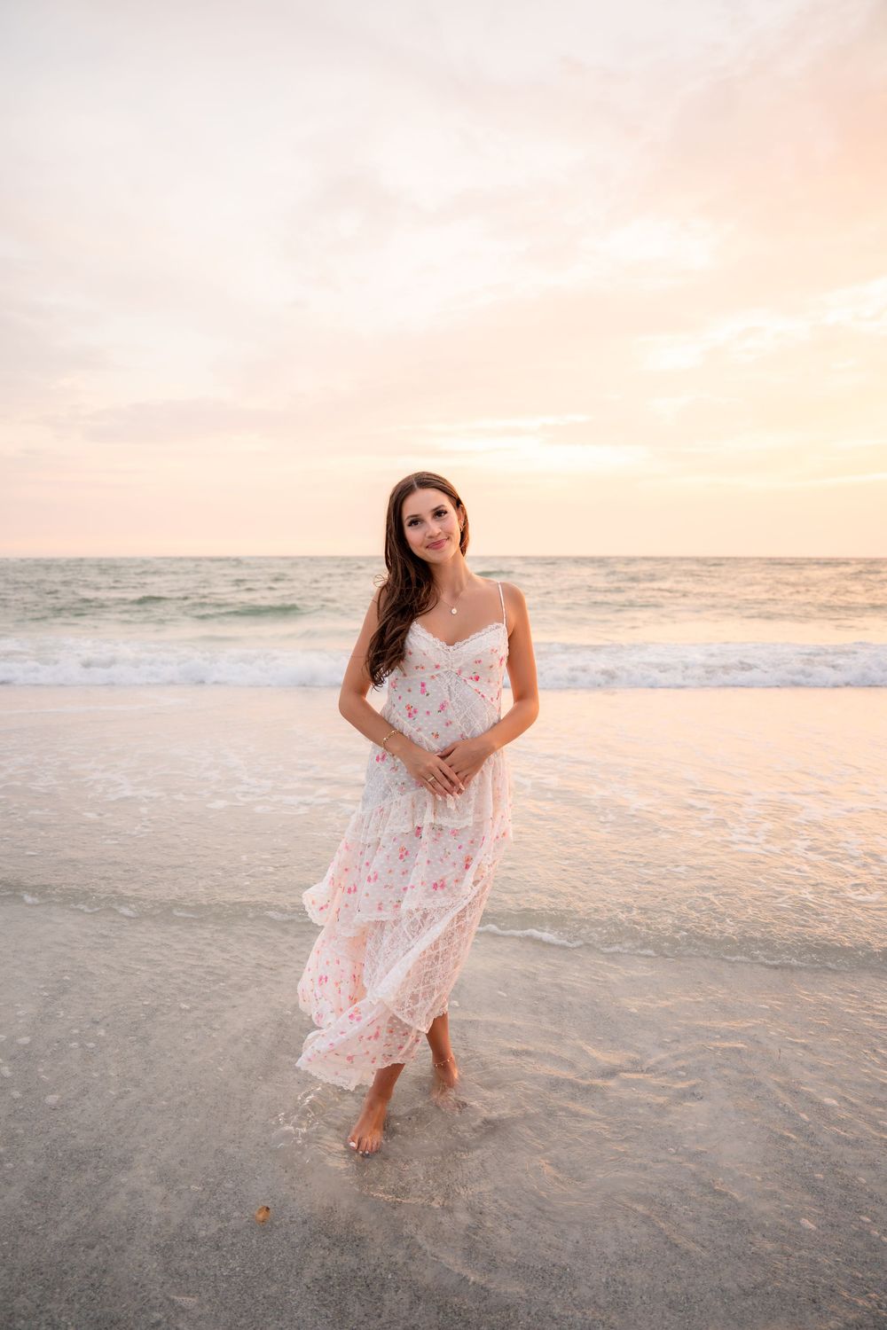 Brunette woman in white floral maxi dress smiling while walking on beach at sunset.