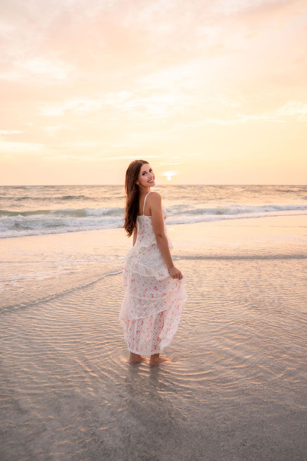 Smiling woman in white floral dress wading in shallow ocean water at sunset.