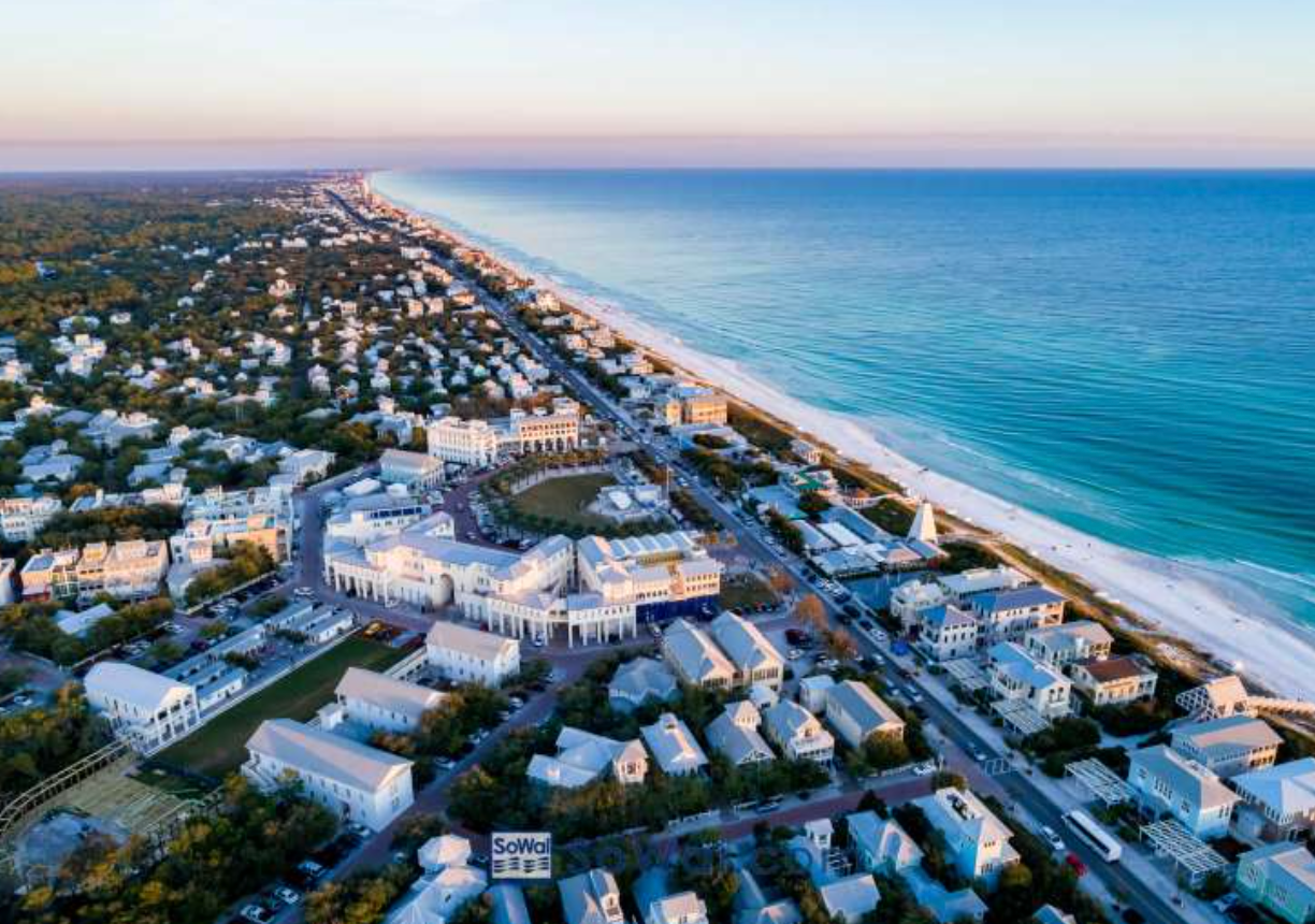 Aerial view of a coastal town with white buildings, lush greenery, and turquoise ocean waters at sunset.