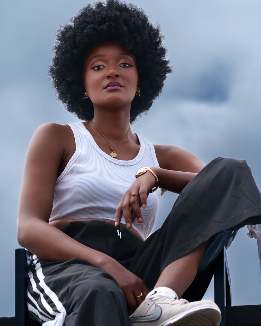 Young Black woman with natural afro hair sitting outdoors in a white tank top and dark track pants.