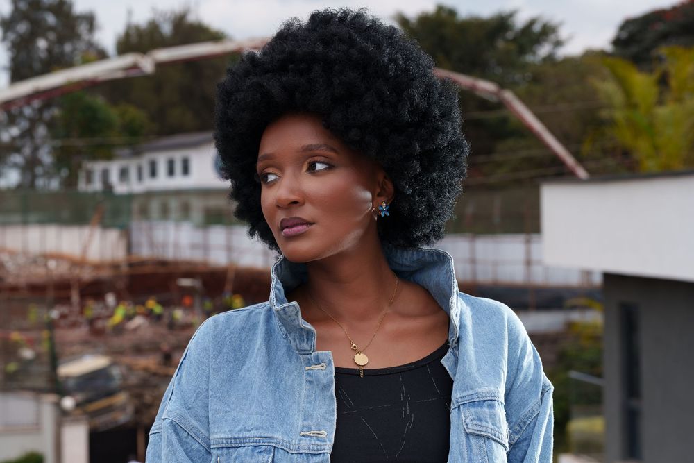 Black woman with large afro hairstyle wearing denim jacket and black top poses outdoors at urban location.