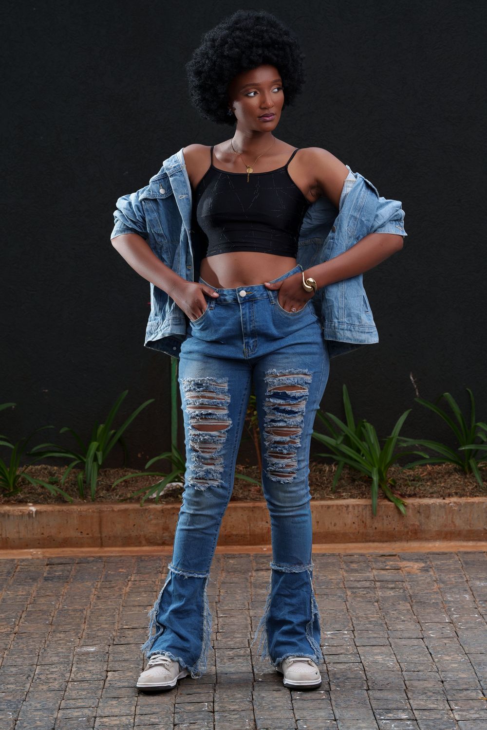 Stylish Black woman with afro stands in ripped flare jeans and denim jacket with black crop top against dark wall.