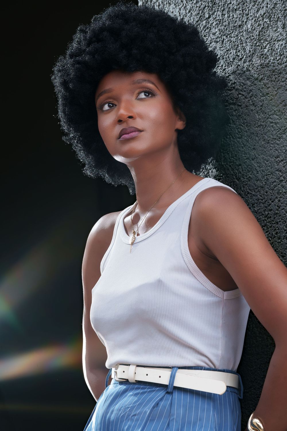 Confident Black woman with afro hairstyle leaning against dark wall, wearing white tank top and blue striped pants.