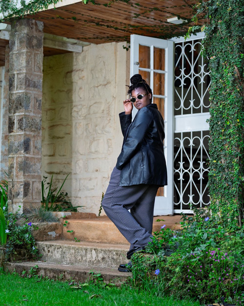 Fashionable Black woman in black leather blazer and patterned pants posing near stone building entrance with greenery.
