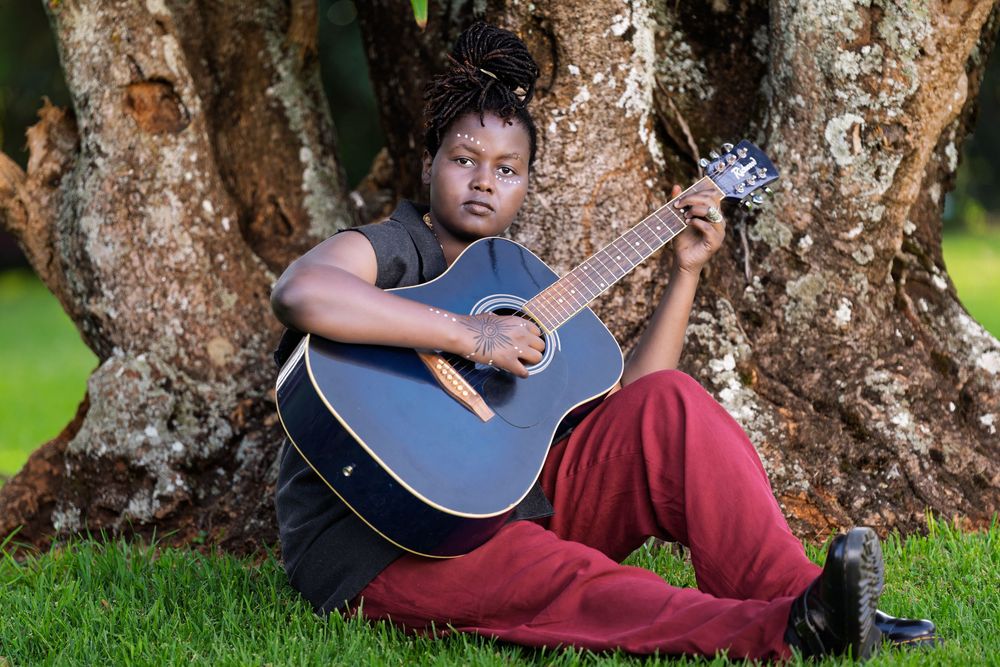 A young woman with locs sits against a large tree trunk holding a blue acoustic guitar in a park.