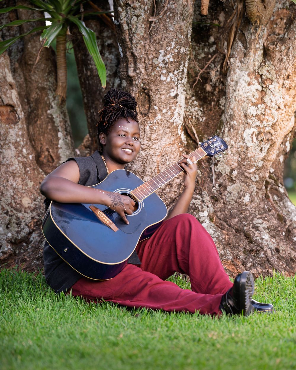 A smiling woman with locs sits on grass against a tree, playing a blue acoustic guitar in red pants.